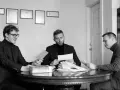 Trzech mężczyzn przy okrągłym stole analizuje dokumenty. Jeden z nich, z okularami, podaje kopertę. W środku siedzi mężczyzna z zarostem, przeglądający stos papierów, a po jego prawej stronie siedzi mężczyzna w kratkę, również z dokumentem. W tle wiszą...