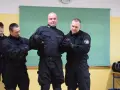 Funkcjonariusze w czarnych mundurach ćwiczą użycie środków przymusu kpk. Jeden z nich jest przytrzymywany przez dwóch kolegów.