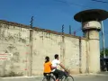Para rowerzystów mija wysoki mur z drutem kolczastym, przypominający więzienie w Tajlandii. Obok stoi wieża strażnicza.