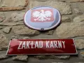 Tablica z napisem "ZAKŁAD KARNY" i godło Polski na kamiennej ścianie.