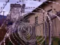 Kolczasty drut tworzy spiralę przed budynkiem. W tle widać góry i przemysłową konstrukcję. Może to miejsce, gdzie zapadały decyzje komisji penitencjarnej.