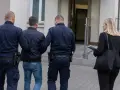 Dwóch policjantów eskortuje mężczyznę, który ma przesłanki do zatrzymania. Obok idzie kobieta z teczką.
