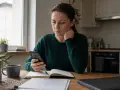 Kobieta zamyślona patrzy na telefon, zastanawiając się, czy grożenie sądem jest karalne. Na stole notatnik i kubek kawy.