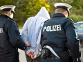 Dwóch policjantów w mundurach z napisem "POLIZEI" prowadzi zatrzymanego mężczyznę w kapturze. Zastanawiasz się, ile trwa areszt w Niemczech?