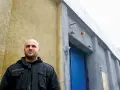 Łysy mężczyzna w czarnej kurtce stoi przed budynkiem z niebieskimi drzwiami i drutem kolczastym. Może to być miejsce, gdzie słychać gwara przestępcza.