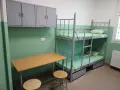 Prosta cela przejściowa z łóżkiem piętrowym, biurkiem i szafkami.