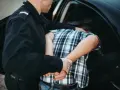 Policjant zakłada kajdanki zatrzymanemu mężczyźnie w kratkę, który nie stawia oporu. To jego dobrowolne poddanie się karze.