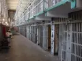 Długi korytarz z rzędami cel, przypominający wnętrze więzienia Riker's Island. Metalowe kraty i drzwi cel tworzą ponury krajobraz.