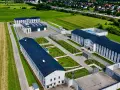 Nowoczesny kompleks budynków otoczony ogrodzeniem, przypominający najnowsze więzienie w Polsce, otoczony polami i domami mieszkalnymi.