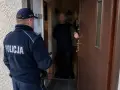 Policjant w mundurze z napisem POLICJA stoi przed drzwiami, gdzie inny mężczyzna otwiera wejście. Sytuacja może być związana z art. 221 kk.