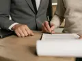 Dwie osoby przy drewnianym stole podpisują dokument. Jedna z nich, ubrana w garnitur, ma na palcu obrączkę. To może być początek postępowania nakazowego.