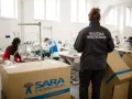Pracownicy wykonują pracę w więzieniu, szyjąc odzież. Nadzorca w kurtce z napisem "Służba Więzienna" obserwuje proces.