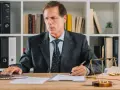 Prawnik w garniturze pracuje przy laptopie, podpisując dokumenty. Na biurku waga sprawiedliwości i księga. To jego upoważnienie dla adwokata.