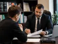 Prawnik w garniturze omawia dokumenty z klientem. Na półkach w tle widać kodeksy. Klient, jak nieświadomy słup, słucha uważnie.