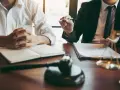 Prawnik omawia z klientem dokumenty, być może dotyczące przepustki z więzienia. Na stole leży młotek sędziowski i waga sprawiedliwości.