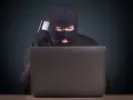 Maskowany mężczyzna z kartą kredytową w ręku, pochylony nad laptopem, ilustruje przykłady przestępstw w sieci.