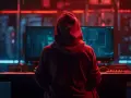 Osoba w czerwonej bluzie z kapturem siedzi przed monitorami, na których widać kod. W tle migoczą czerwone i niebieskie światła. To może być scena z filmu o hakerach, gdzie 130 kk to być może liczba danych lub zaszyfrowanych informacji.