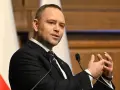 Mężczyzna w garniturze przemawia, gestykulując rękami. W tle biało-czerwona flaga, symbol prawa i sprawiedliwości.