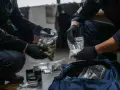 Policjanci w rękawiczkach pokazują zabezpieczoną znaczną ilość narkotyków w woreczkach. W tle torba i inne pojemniki.