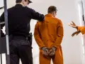 Strażnik eskortuje skutego mężczyznę w pomarańczowym kombinezonie. To jak wygląda przyjęcie do więzienia.