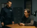 Policjant w mundurze stoi nad mężczyzną siedzącym przy biurku w pokoju przesłuchań. Mężczyzna wygląda na zamyślonego, a policjant na niego patrzy. To typowy dołek policyjny.