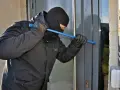 Osoba w kominiarce i rękawiczkach próbuje wyważyć drzwi łomem. To przykład kradzieży z włamaniem.