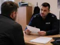 Policjant rozmawia z mężczyzną, trzymając dokumenty. Prawdopodobnie dotyczy to wniosku o zmianę miejsca dozoru policyjnego.