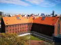 Widok na ceglany budynek z pomarańczowym dachem, otoczony ogrodzeniem z drutu kolczastego. W tle miejski krajobraz z wieżowcami. To zakład karny Służewiec.