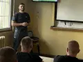 Strażnik więzienny prowadzi szkolenie w sali przypominającej izolatkę. Na ekranie prezentacja, a przed nim słuchacze.