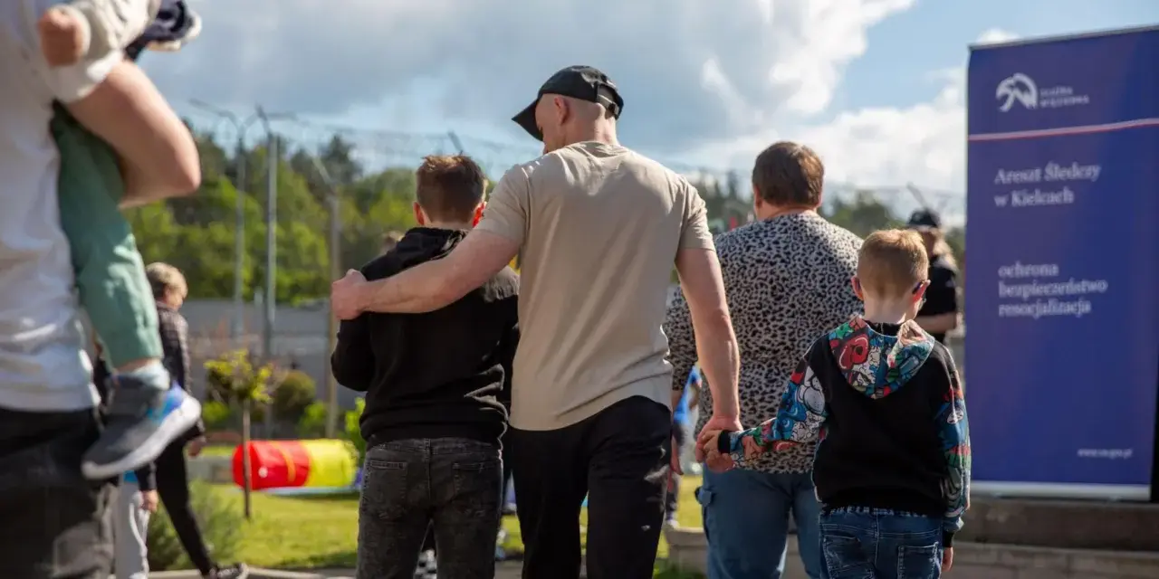 Rodzina idzie w stronę aresztu. Dzieci trzymają się za ręce, a dorośli obejmują się nawzajem. W tle widać ogrodzenie i tablicę informacyjną. To obraz matki za kratami, która spotyka się z bliskimi.