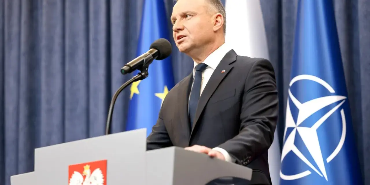 Prezydent Andrzej Duda przemawia przy mównicy z godłem Polski. Za nim flagi UE i NATO. Może to być związane z ułaskawieniem prezydenta.