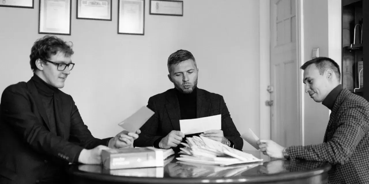 Trzech mężczyzn przy okrągłym stole analizuje dokumenty. Jeden z nich, z okularami, podaje kopertę. W środku siedzi mężczyzna z zarostem, przeglądający stos papierów, a po jego prawej stronie siedzi mężczyzna w kratkę, również z dokumentem. W tle wiszą...