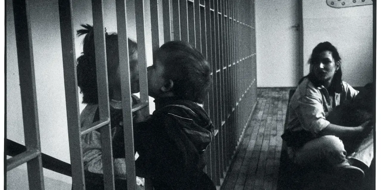 Dzieci przy kratach, zastanawiamy się, czy zabierać dziecko do zakładu karnego. Kobieta siedzi w tle.
