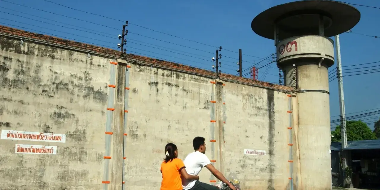 Para rowerzystów mija wysoki mur z drutem kolczastym, przypominający więzienie w Tajlandii. Obok stoi wieża strażnicza.