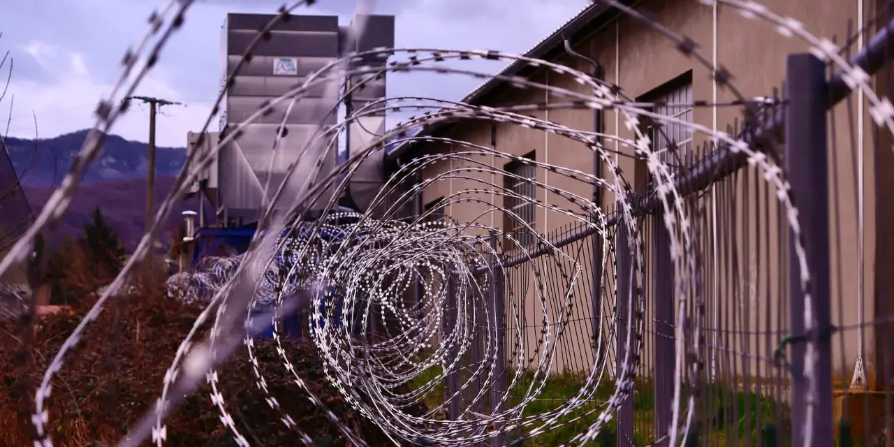 Kolczasty drut tworzy spiralę przed budynkiem. W tle widać góry i przemysłową konstrukcję. Może to miejsce, gdzie zapadały decyzje komisji penitencjarnej.