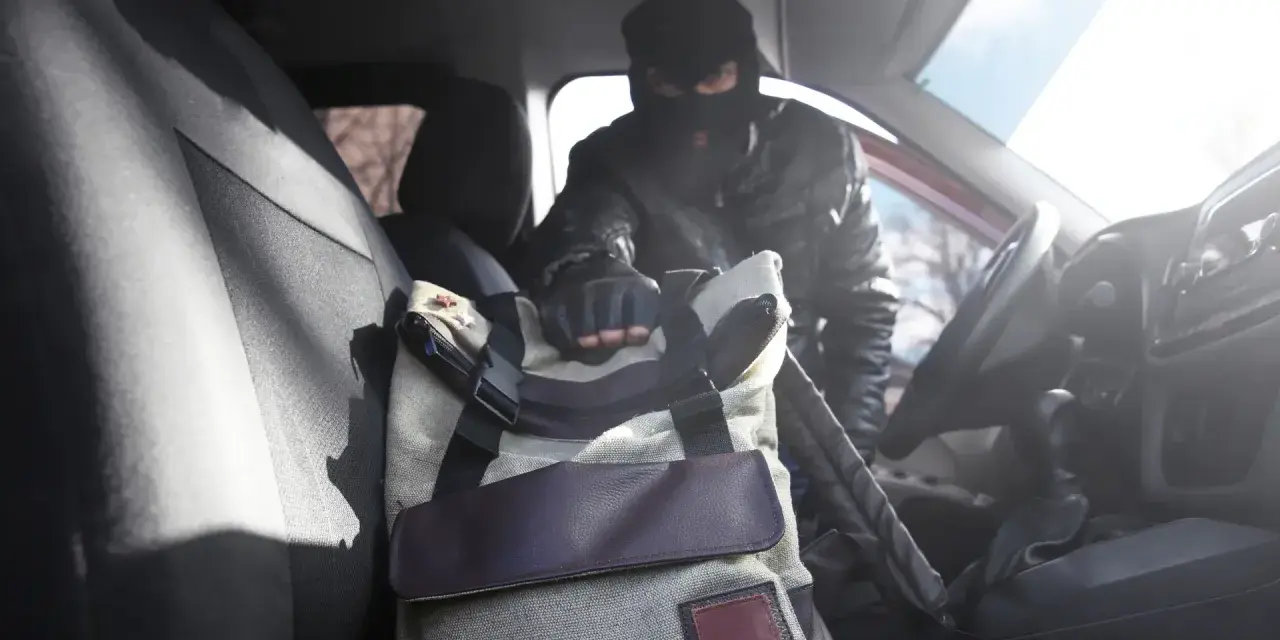 Złodziej w masce sięga po torbę w samochodzie. To jeden z rodzajów kradzieży, które mogą spotkać każdego.