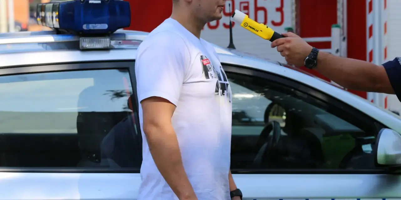 Policjant przeprowadza procedurę badania alkomatem. Mężczyzna dmucha w urządzenie przy radiowozie.