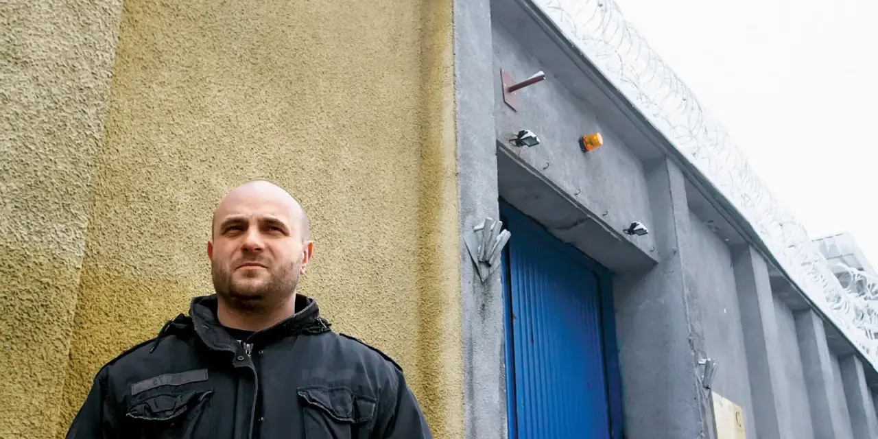 Łysy mężczyzna w czarnej kurtce stoi przed budynkiem z niebieskimi drzwiami i drutem kolczastym. Może to być miejsce, gdzie słychać gwara przestępcza.