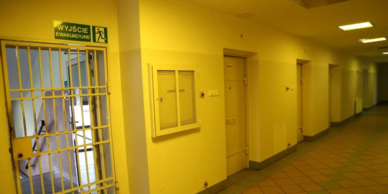 Korytarz więzienny z kratami i drzwiami cel. Nad drzwiami znak "Wyjście ewakuacyjne".