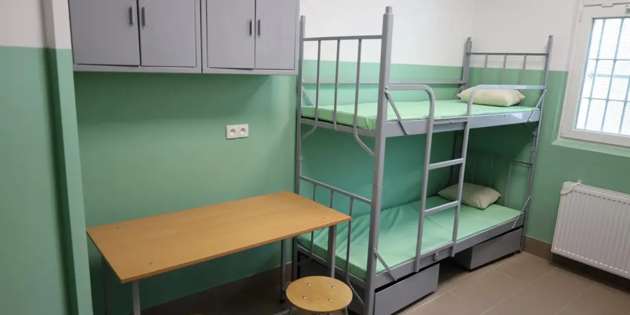 Prosta cela przejściowa z łóżkiem piętrowym, biurkiem i szafkami.