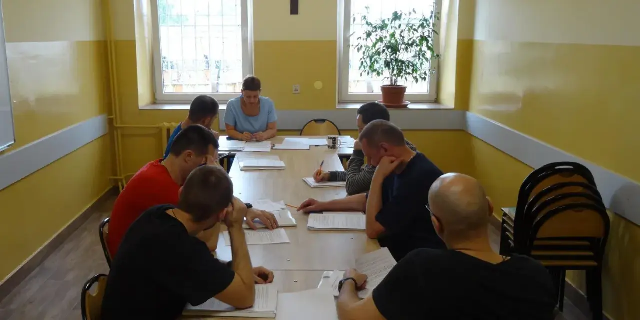 Grupa mężczyzn uczestniczy w zajęciach, które mogą być częścią system programowanego oddziaływania. Siedzą przy stole, studiując dokumenty.