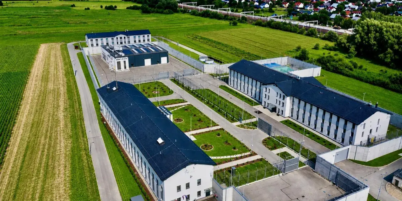 Nowoczesny kompleks budynków otoczony ogrodzeniem, przypominający najnowsze więzienie w Polsce, otoczony polami i domami mieszkalnymi.