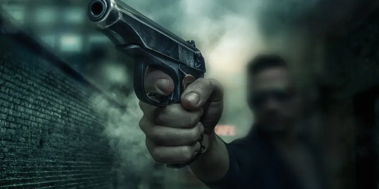 Dłoń trzyma pistolet, gotowa do strzału. Scena sugeruje sytuację z art. 64 par. 2 kk, gdzie groźba użycia broni może być kluczowa.