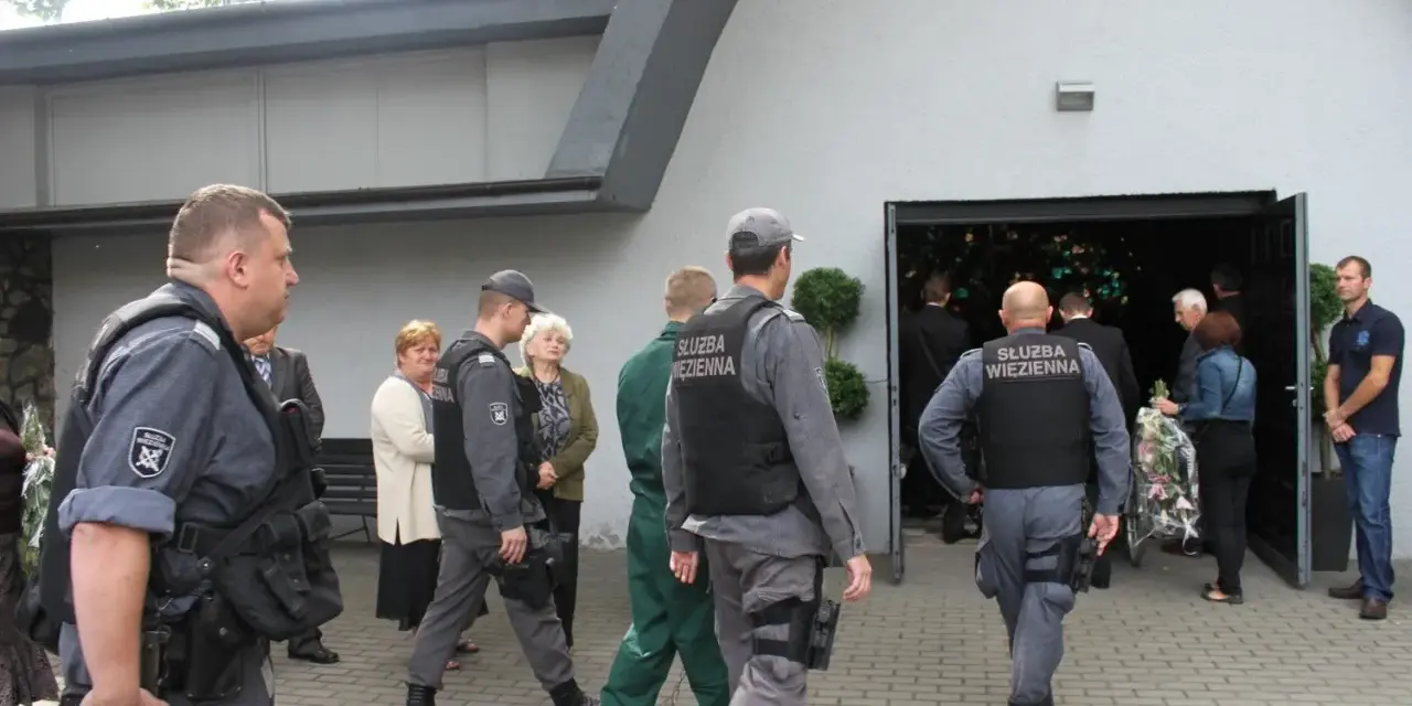 Strażnicy więzienni eskortują więźnia na pogrzeb. Widać ich w mundurach przed budynkiem, gdzie odbywa się ceremonia.