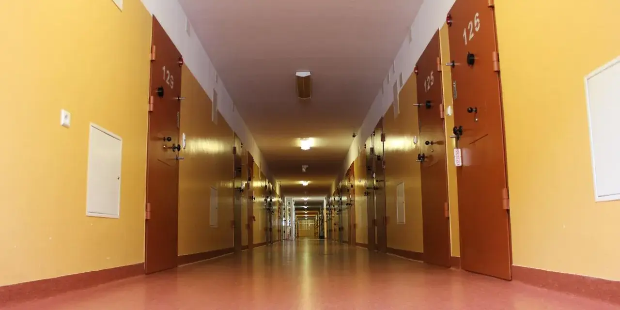 Długi korytarz z rzędem drzwi, typowy dla zakładu karnego dla kobiet. Jasne ściany i drzwi z numerami cel.
