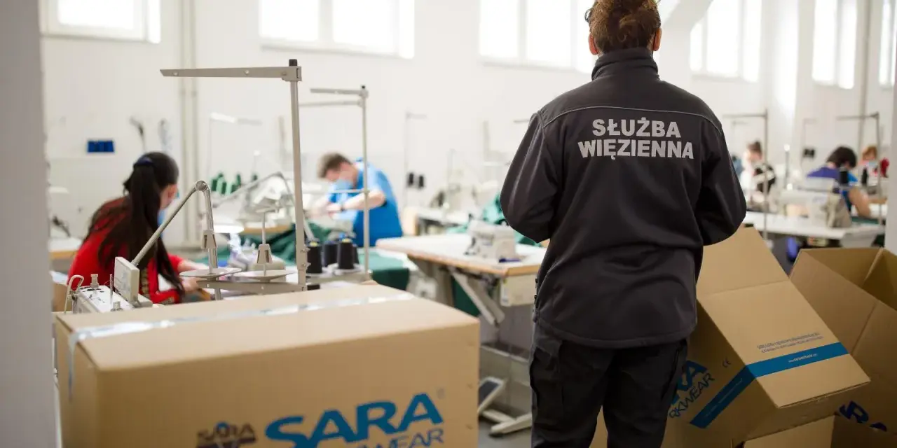 Pracownicy wykonują pracę w więzieniu, szyjąc odzież. Nadzorca w kurtce z napisem "Służba Więzienna" obserwuje proces.