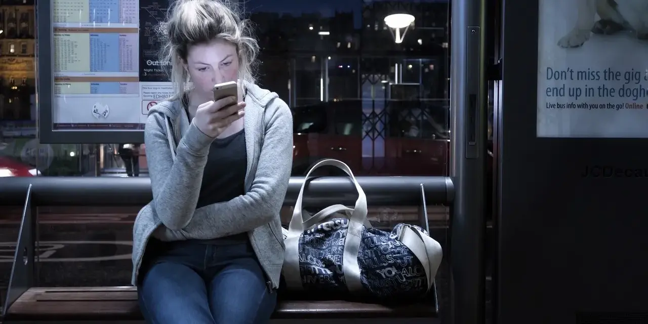 Dziewczyna na przystanku nocnego autobusu, pochłonięta telefonem, ignoruje uporczywe niepokojenie. Obok torba.