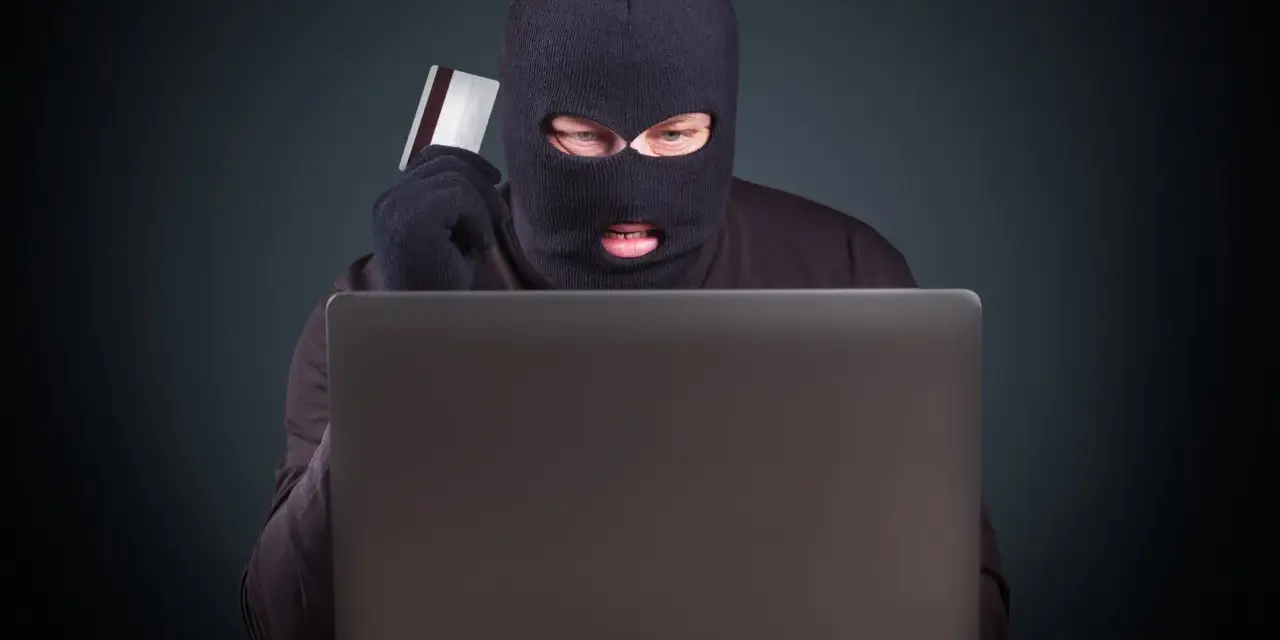 Maskowany mężczyzna z kartą kredytową w ręku, pochylony nad laptopem, ilustruje przykłady przestępstw w sieci.