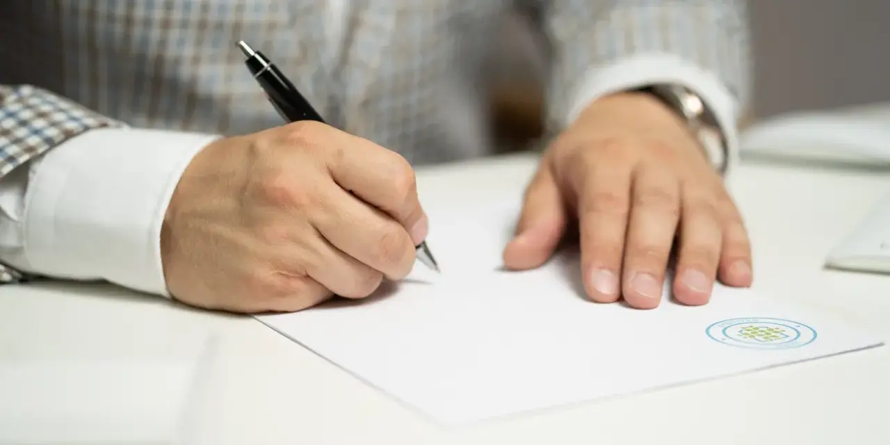 Mężczyzna w eleganckim garniturze podpisuje ważny dokument. Jego ręka pewnie trzyma długopis, a na papierze widnieje logo firmy.