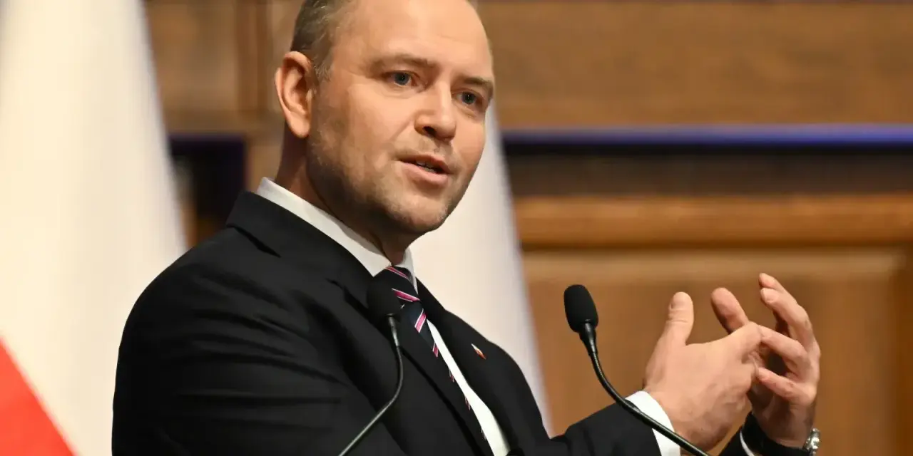 Mężczyzna w garniturze przemawia, gestykulując rękami. W tle biało-czerwona flaga, symbol prawa i sprawiedliwości.